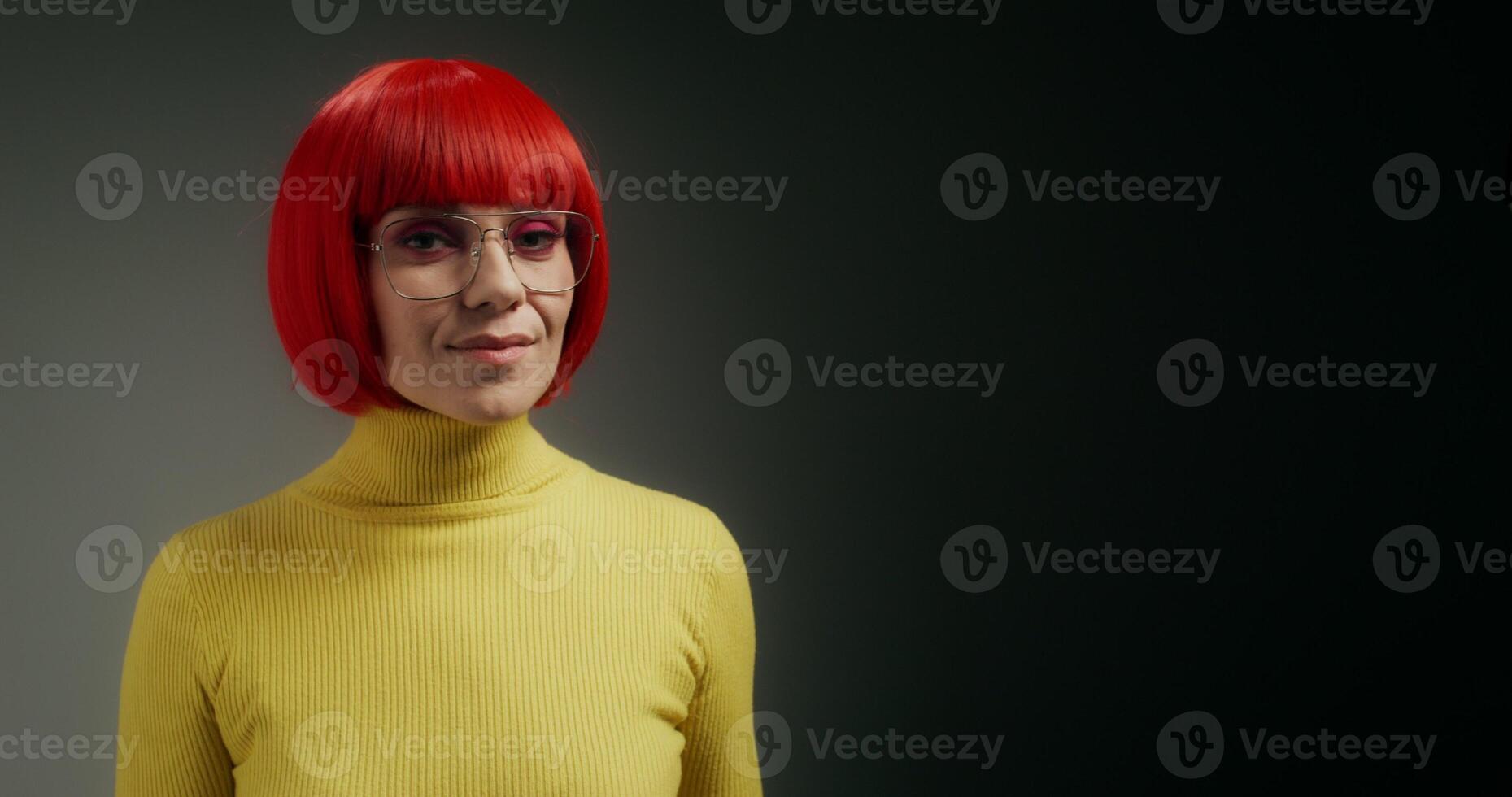 A woman with glasses and an orange wig smiles at the camera, holding a mobile phone with green screen in her hand, standing on a gray background in the studio photo