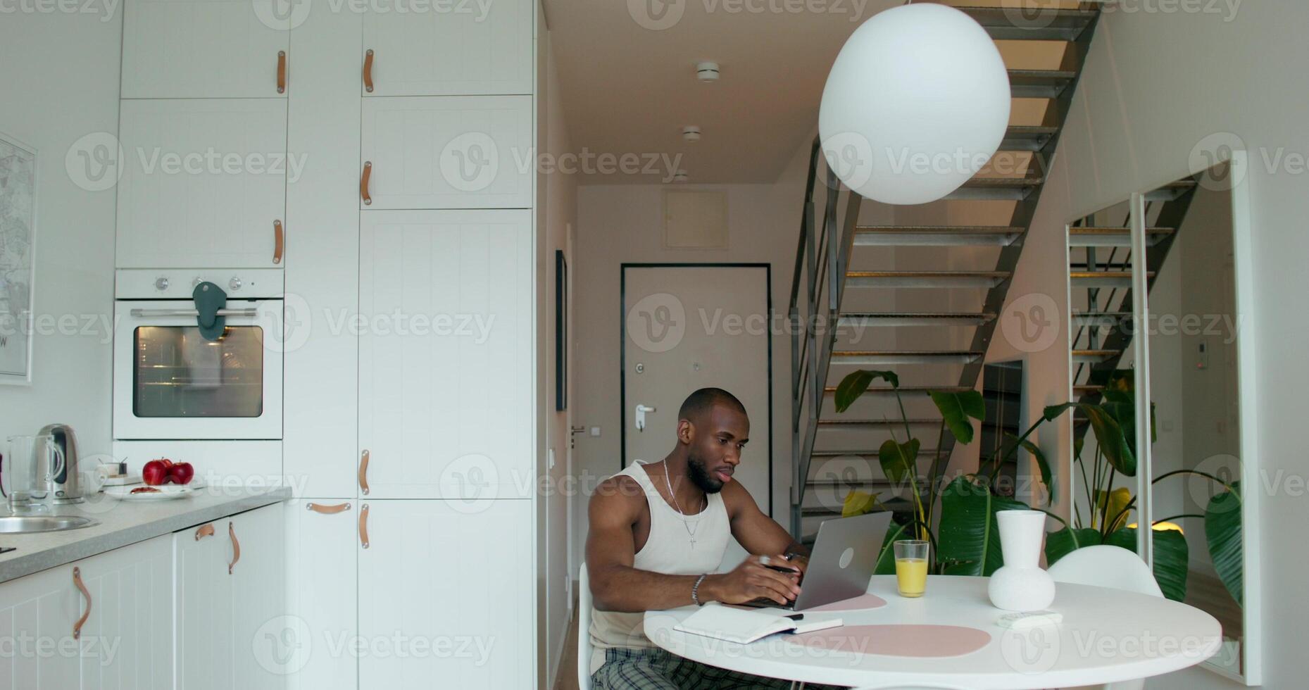 A man in pajamas uses a laptop and writes in a notebook sitting at a table in the kitchen at home with a serious look - kitchen photo