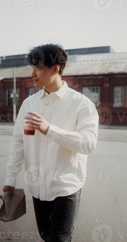 A young man with a paper bag in his hands drinks juice from a disposable cup with a straw while walking around the city - around photo