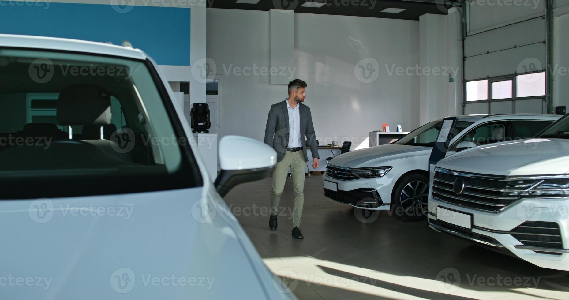 A stylish man chooses a car at a car dealership. He walks between the cars and examines them with a serious look - examines photo