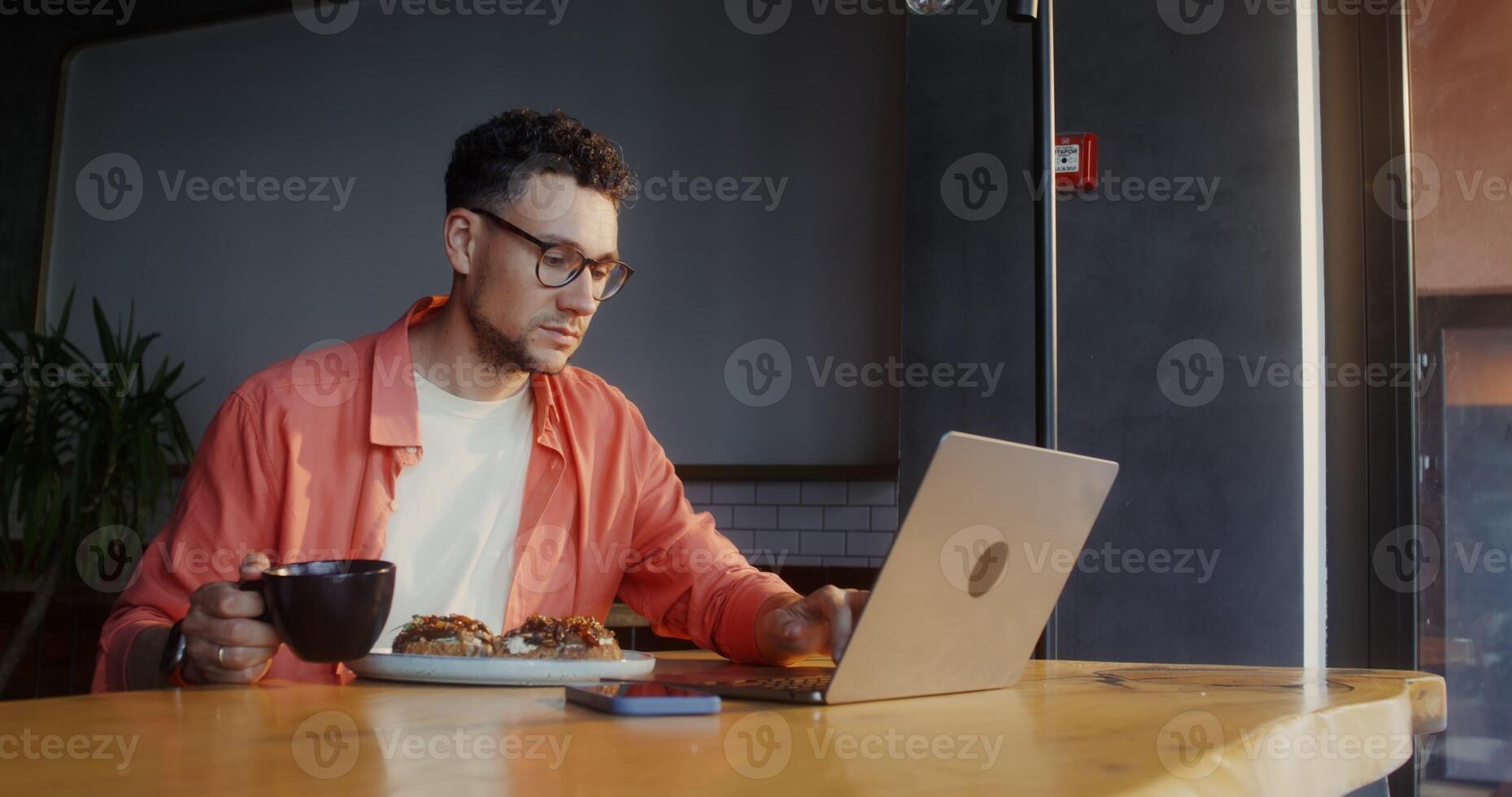 A stylish young man with glasses uses a laptop and drinks coffee sitting at a table in a cafe during vacation photo