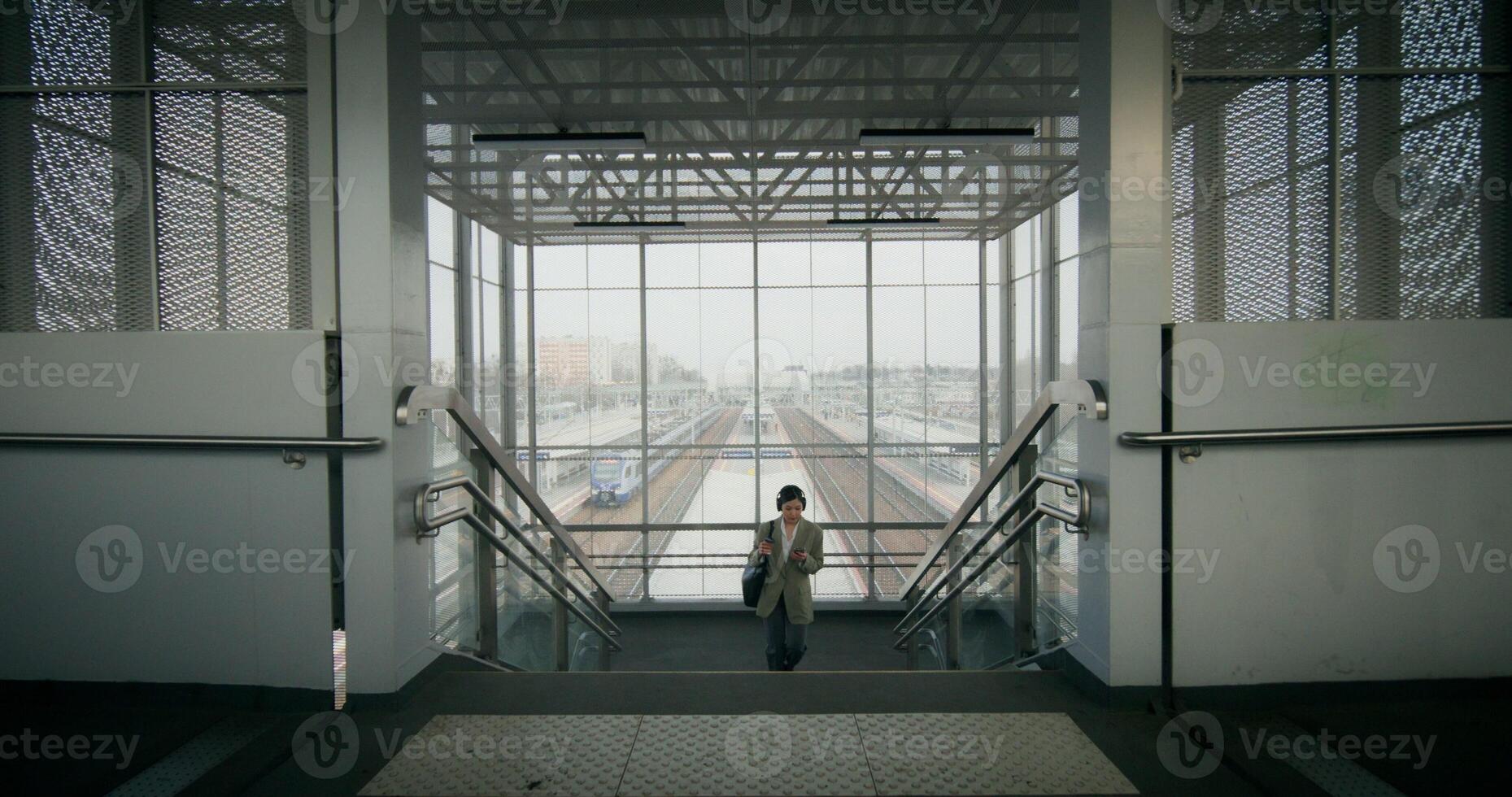 A woman in formal clothes and with headphones on her head uses a mobile phone and drinks coffee from a disposable cup walking along the railway elevated passage photo