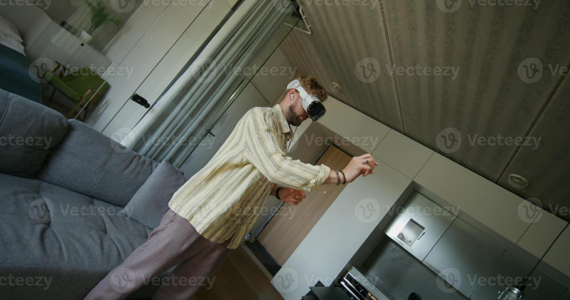 A man in a virtual reality headset and with earbuds in his ears moves his hands standing in a home interior. Daylight photo