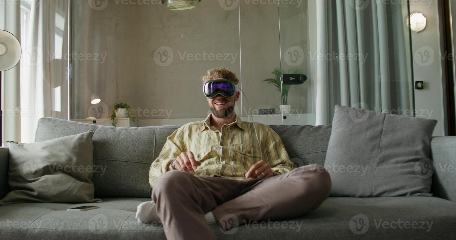 A man in a virtual reality headset puts on earphones and starts a chat sitting on the couch at home. He smiles, talks and moves his arms. Daylight - headset photo