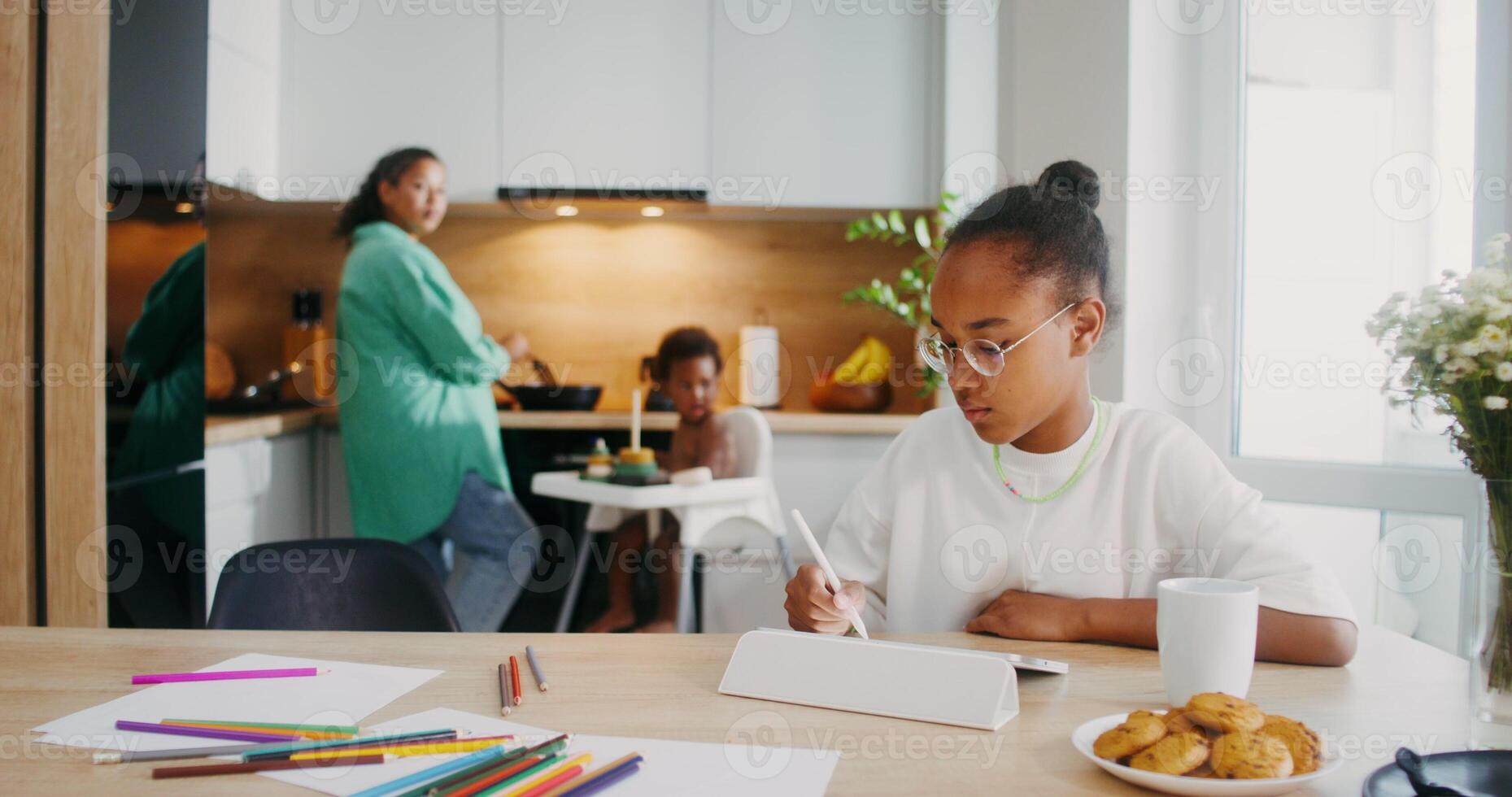 A girl draws with a stylus on a tablet while sitting at the kitchen table at home. Her family in the background is out of focus - tablet photo