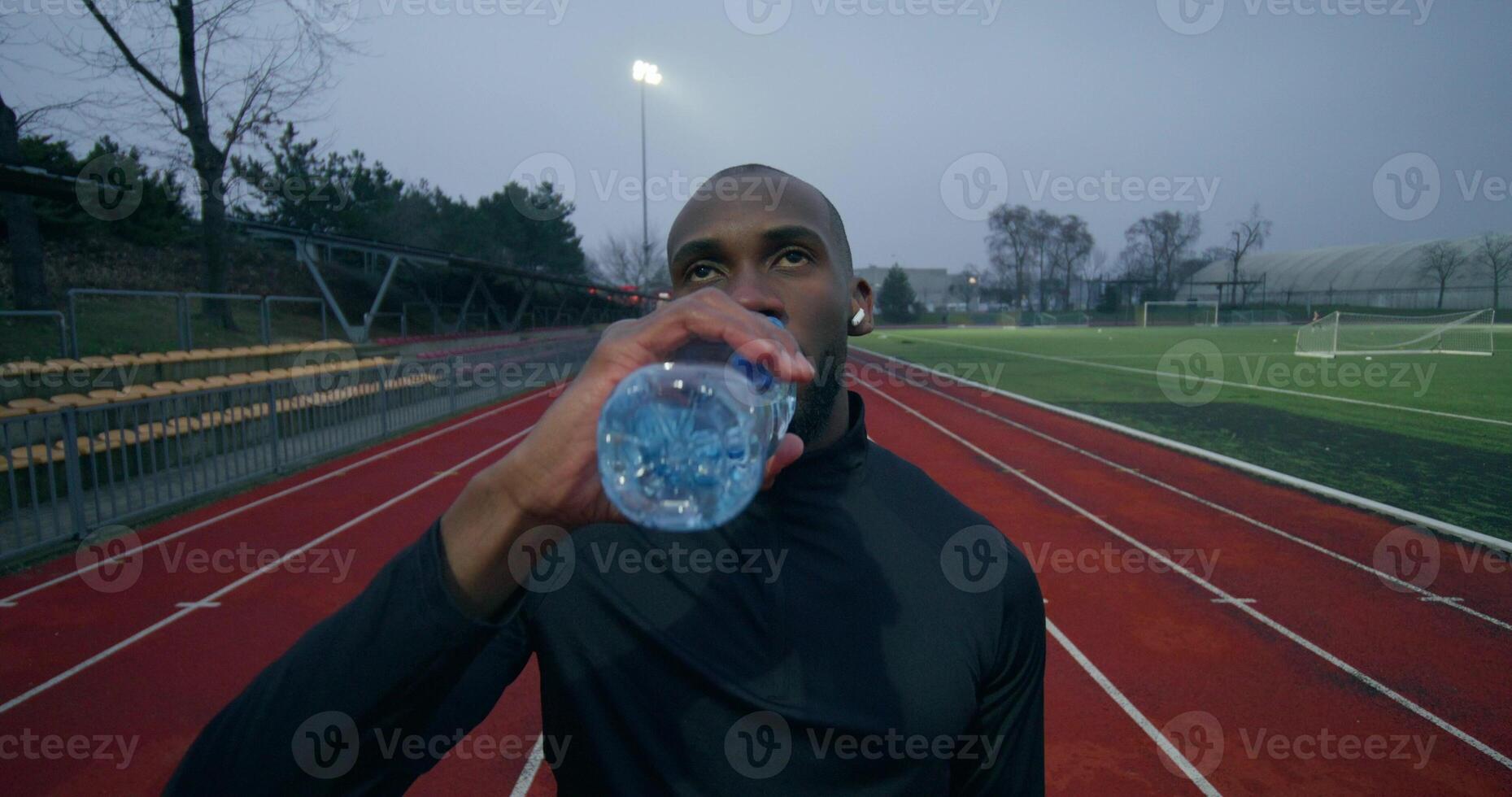 A man in sports clothes, with earphones in his ears, drinks water from a bottle standing at the stadium after jogging photo