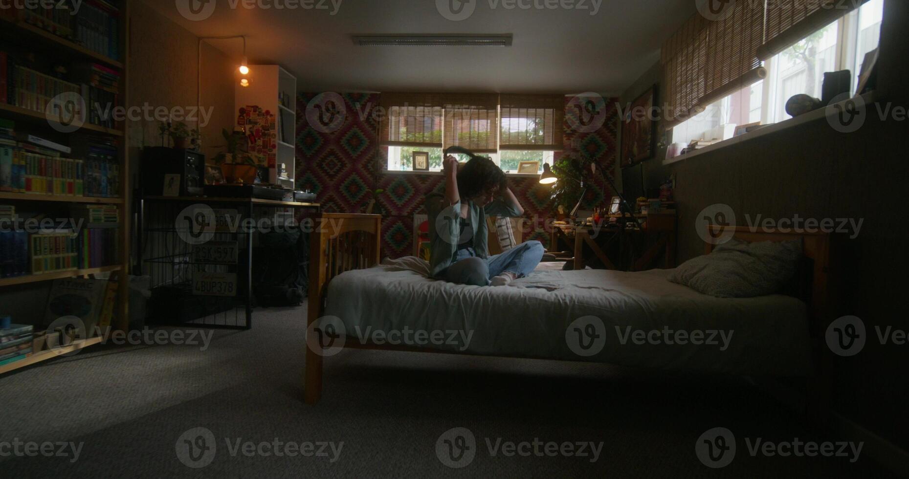 A young woman puts on headphones and uses a mobile phone sitting on the bed. She is upset or depressed. Teenage home interior photo