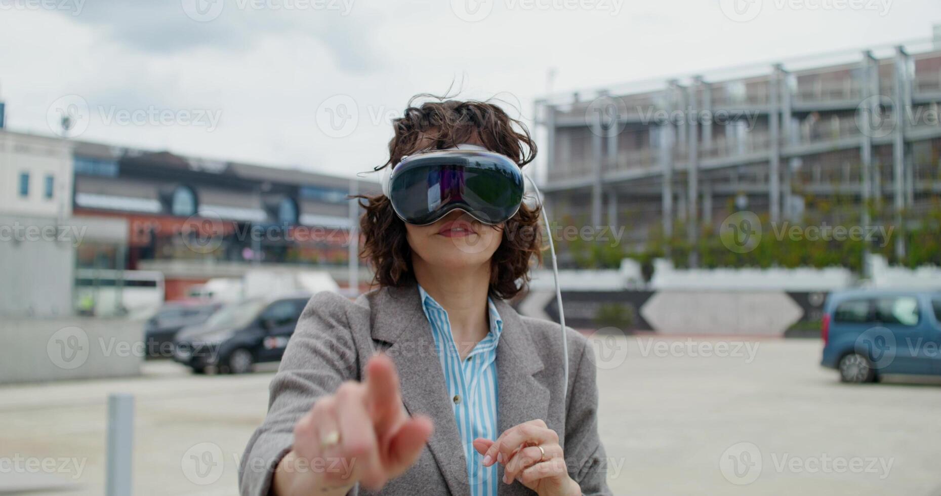A stylish woman in a jacket wearing a virtual reality headset stands on a city street and moves her arms. Summer day - virtual photo