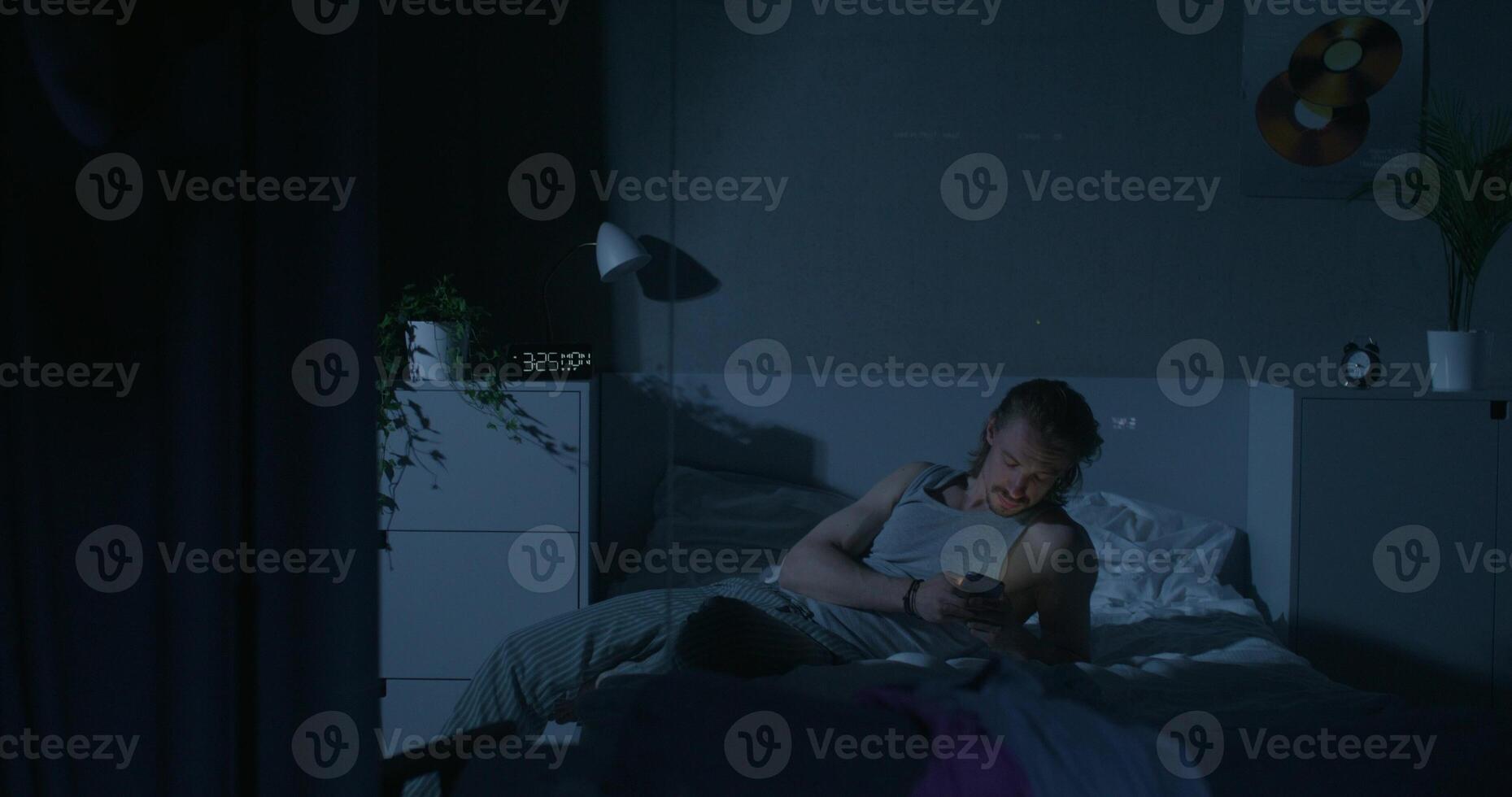 A man in pajama pants uses a mobile phone while sitting on the bed at night. Dark room, home interior - pajama photo