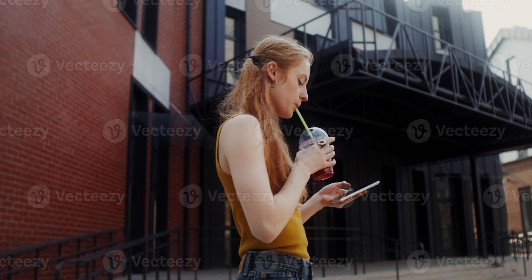 A young red-haired woman uses a mobile phone and drinks juice from a disposable cup with a straw while walking on a city street photo