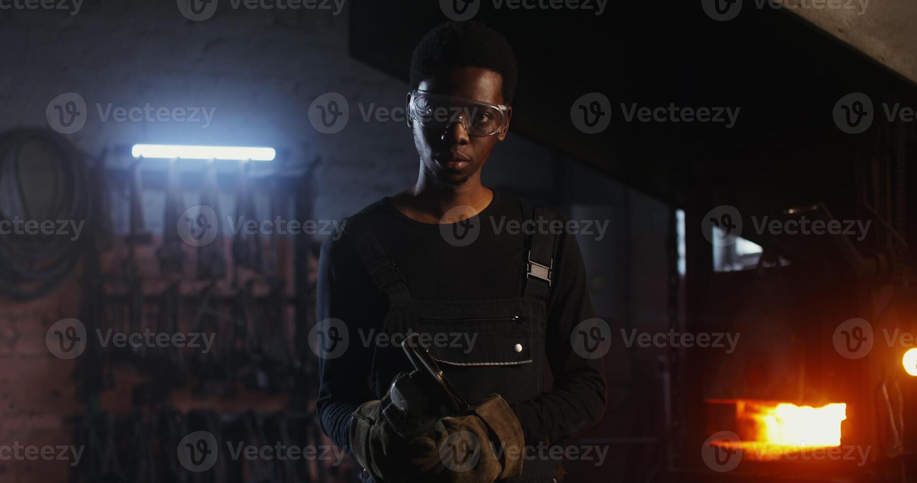 A young blacksmith of African American appearance in goggles and an apron turns on an angle grinder and begins to cut off a piece of metal workpiece while working in a forge. 4k, red komodo - American photo