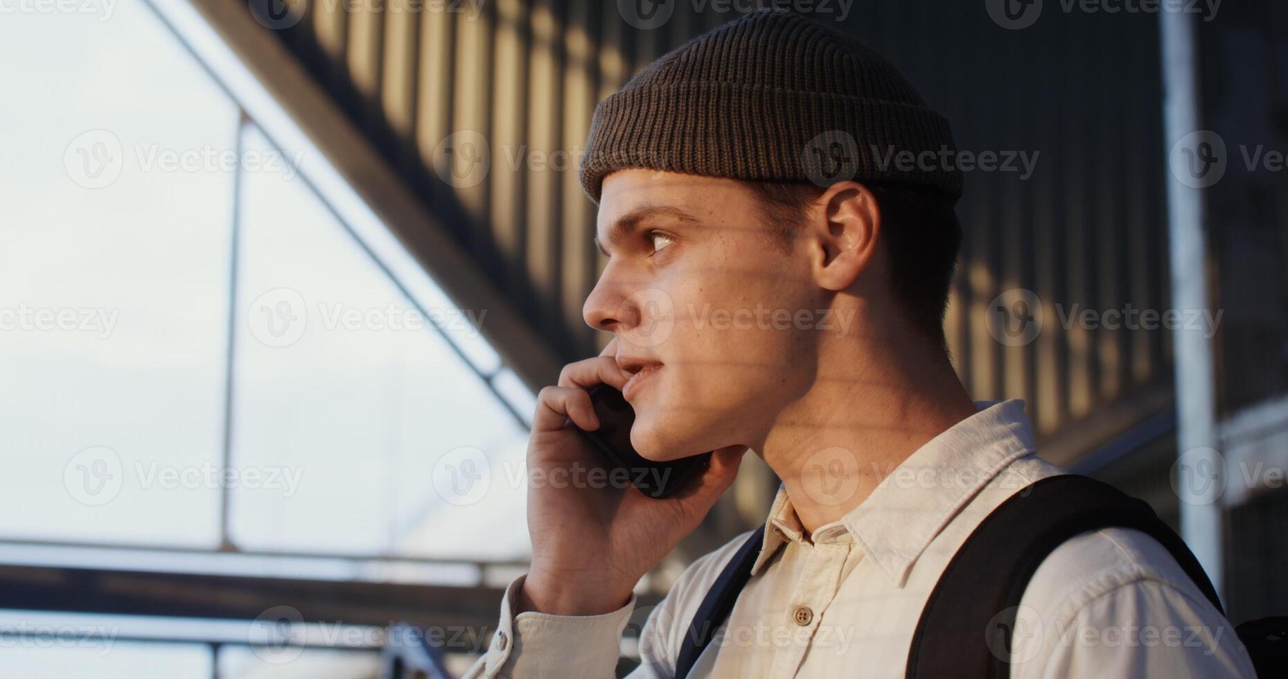 A young man of European appearance, in a knitted hat with a backpack on his back, is talking on the phone, laughing cheerfully - backpack photo
