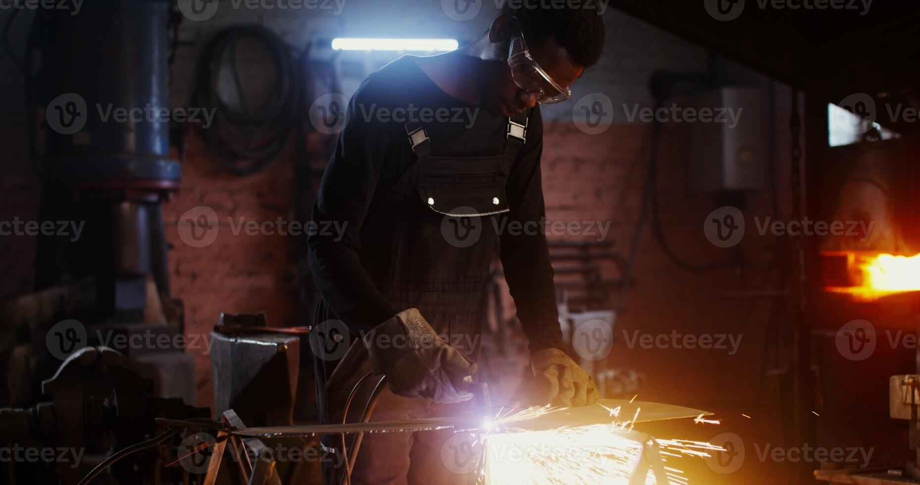 A man of African American appearance, dressed in work clothes and goggles with a concentrated look, uses a plasma cutter, cutting a sheet of iron, while working in a forge. 4k, red komodo - clothes photo