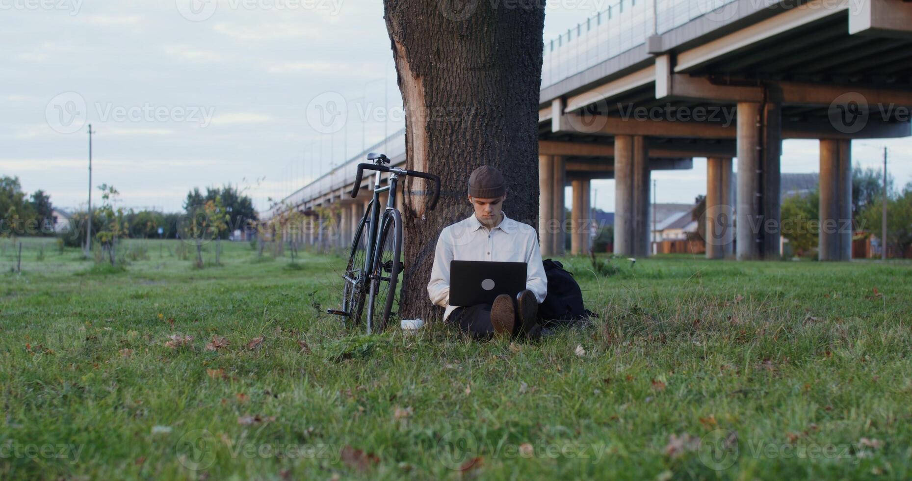 A young man of European appearance, in a knitted hat, uses a laptop and drinks coffee, sitting on the ground under a tree next to a bicycle. photo