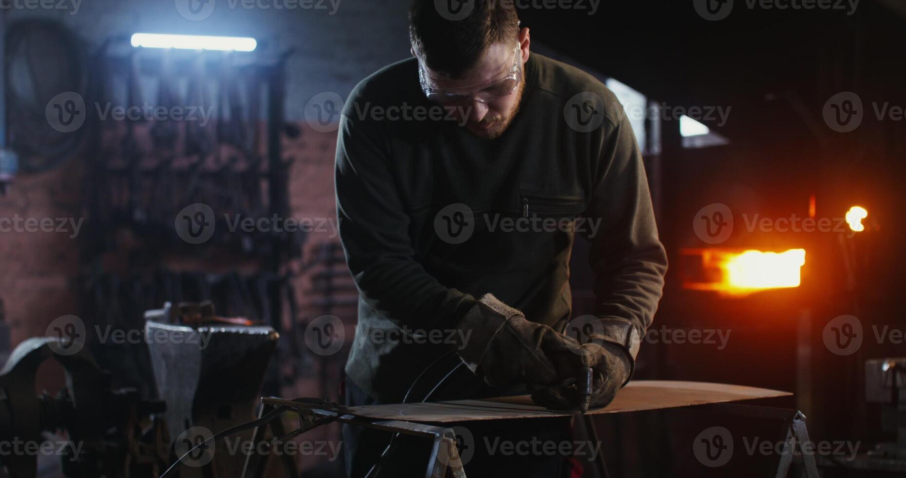 A young man of European appearance, dressed in work clothes and goggles, cuts a sheet of iron with a plasma cutter while working in a forge. 4k, red komodo photo