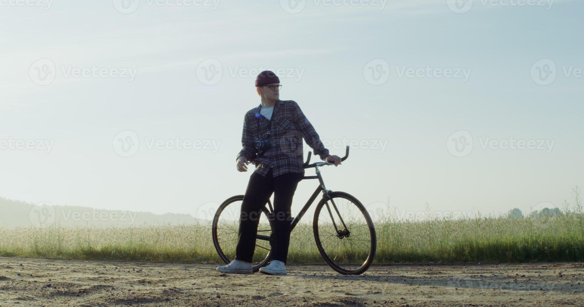 A young man with a camera around his neck stopped near a wheat field, resting after cycling and admiring the beauty of nature in the rays of the rising sun. 4k , red komodo - stopped photo