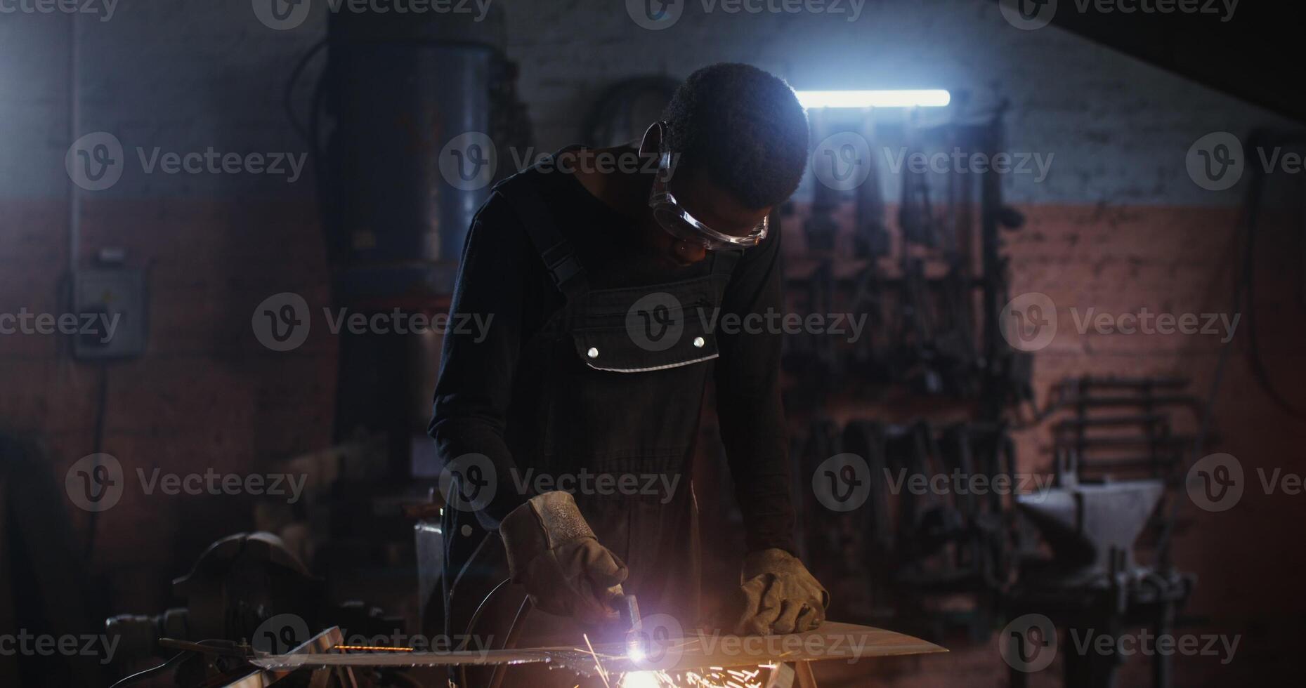 A man of African American appearance, dressed in work clothes and goggles with a concentrated look, uses a plasma cutter, cutting a sheet of iron, while working in a forge. 4k, red komodo - dressed photo