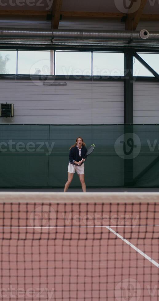 Beautiful young woman playing tennis, hitting the ball with a tennis racket and bouncing during strikes, playing tennis on an indoor tennis court, vertical photo