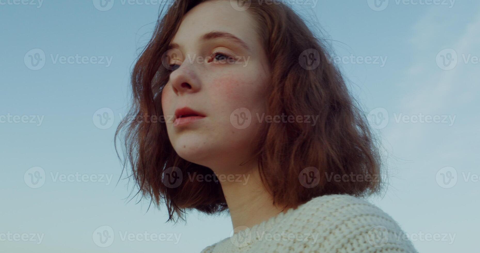 Close-up of the face of a young red-haired woman with a short haircut, looking at the camera and up with a serious look, standing against a blue sky. 4k , red komodo. photo