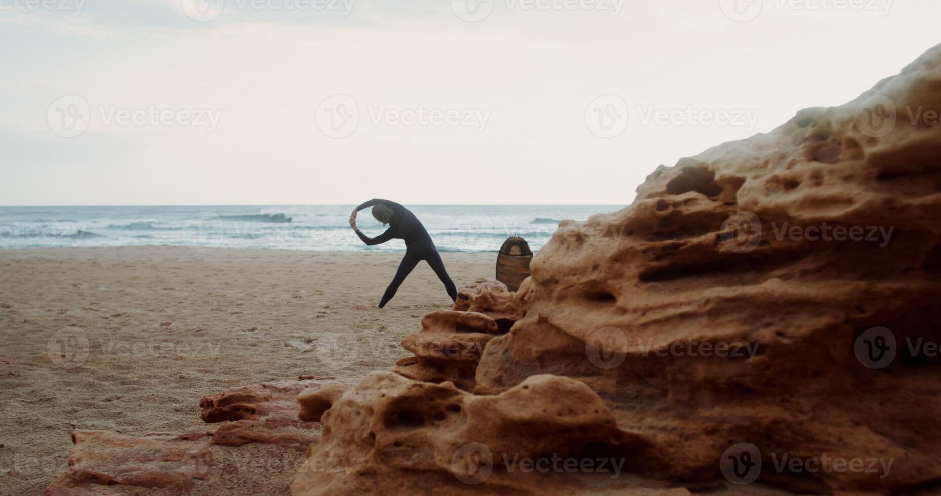 A man in a diving suit does exercises standing on the sandy seashore next to a surfboard stuck in the sand. Rear view - standing photo