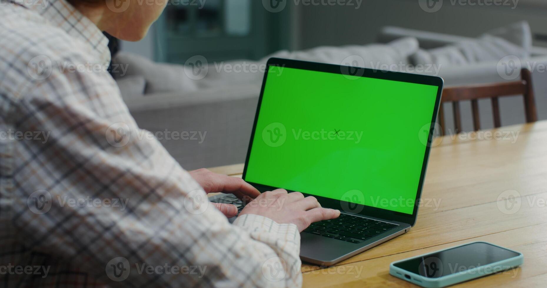 A woman in casual clothes uses a laptop with a green screen while sitting at a table indoors. from behind a woman's shoulder. Copy space, mockup photo