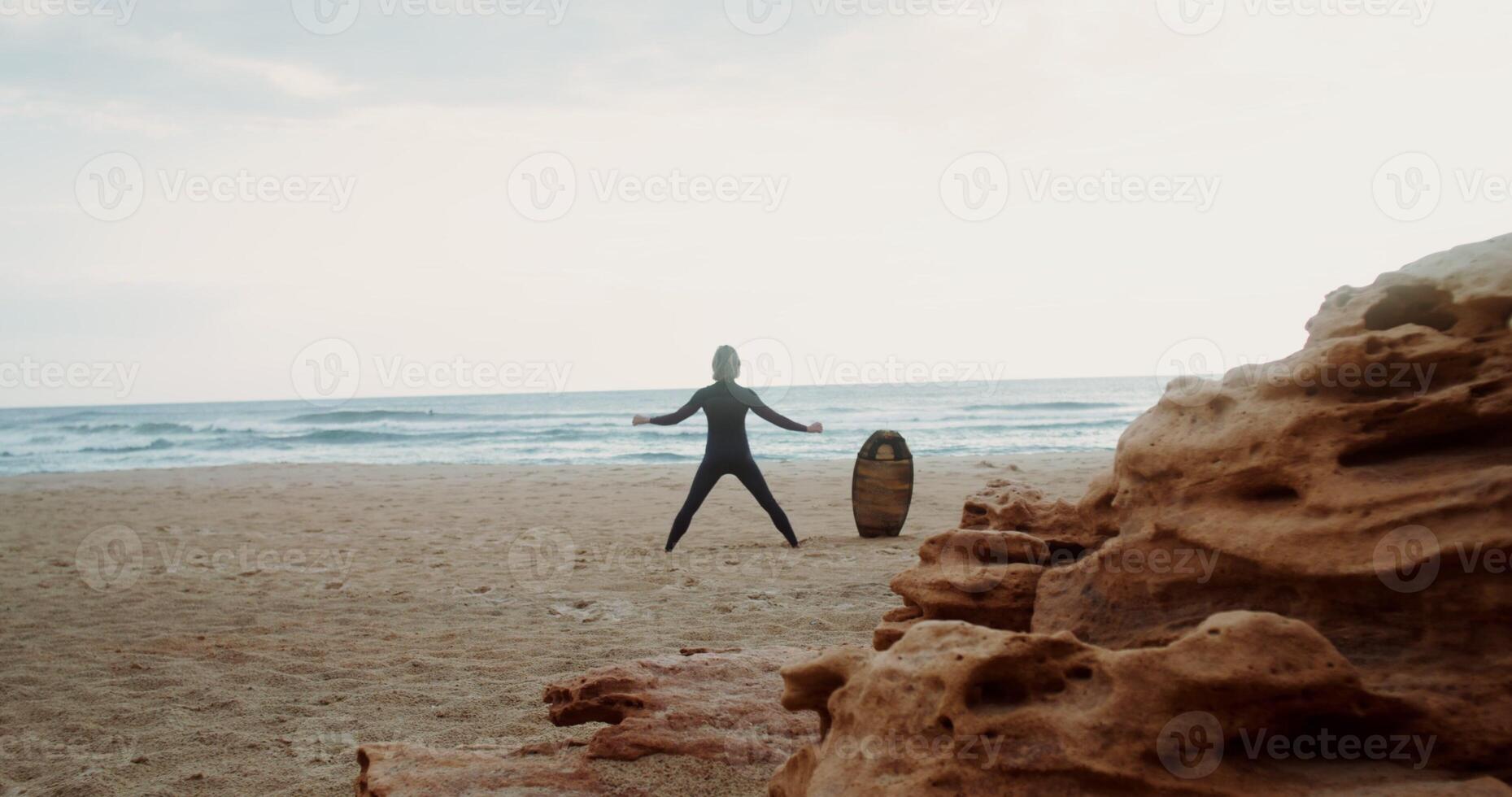 A man in a diving suit does exercises standing on the sandy seashore next to a surfboard stuck in the sand. Rear view photo