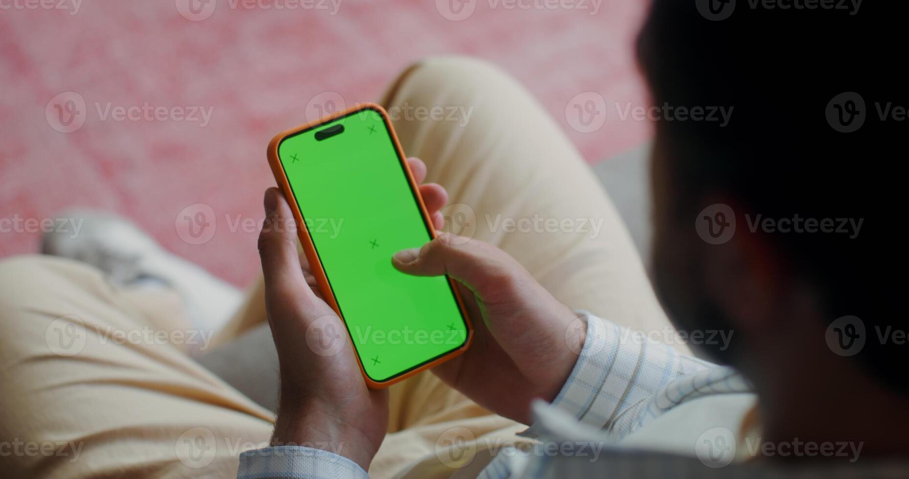 A man in casual clothes scroll the green screen of a mobile phone while sitting on the couch at home. Top view, no face, close-up of a man's hands photo