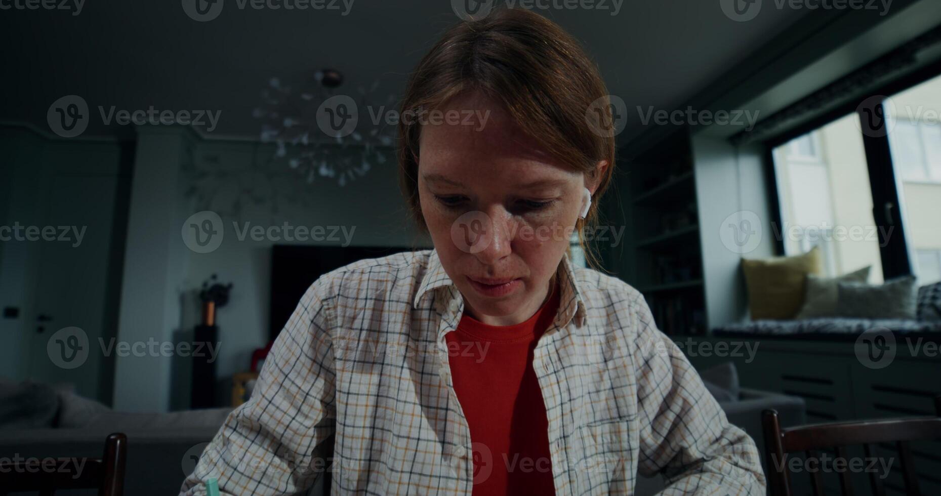 A young woman in casual clothes makes notes in a notebook, and uses a laptop while sitting at a table in a stylish home interior with a serious look photo