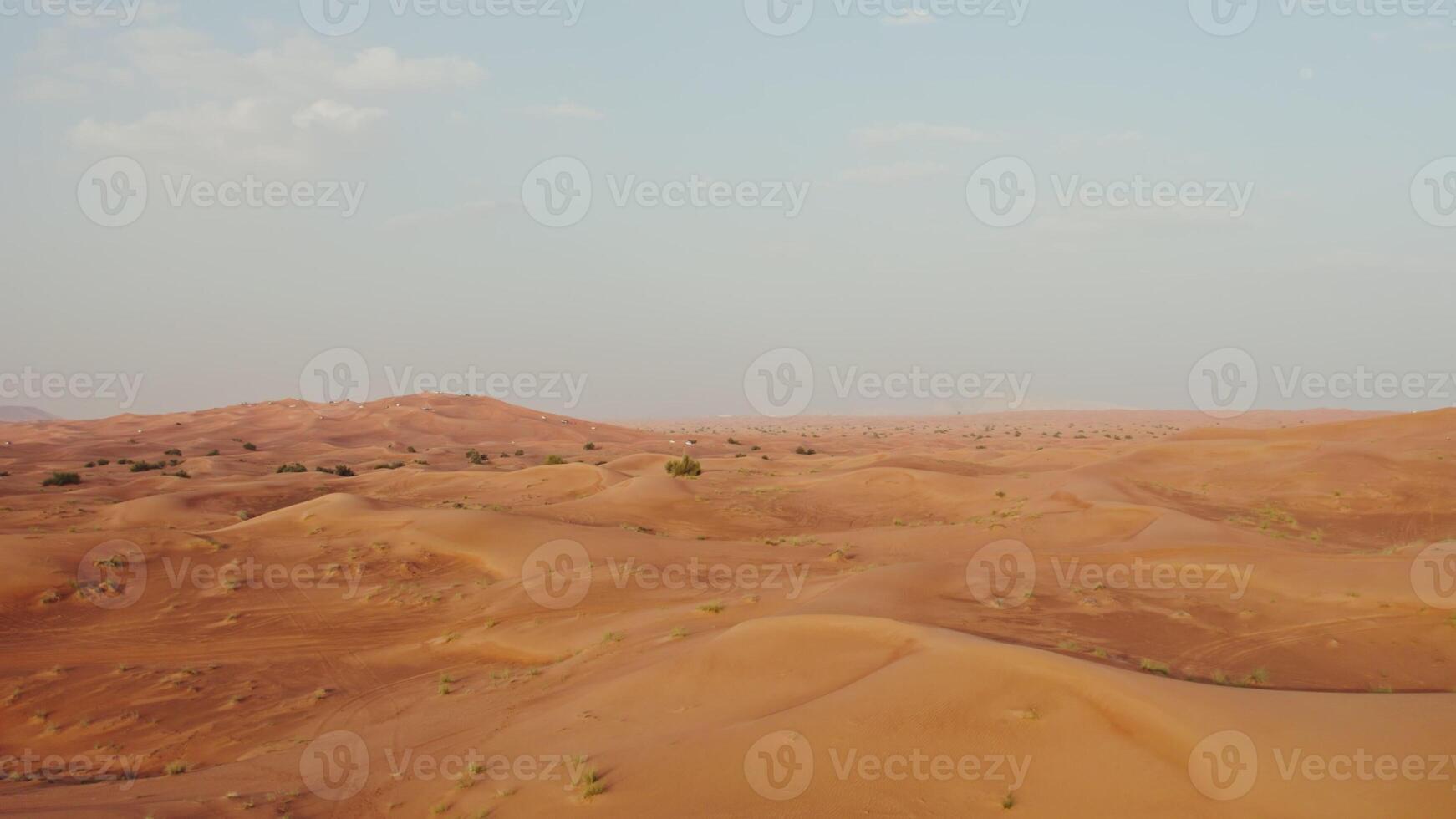 Drone view of a lifeless desert with sparse vegetation among red sand. Cars drive into a sandy mountain in the background - sparse photo