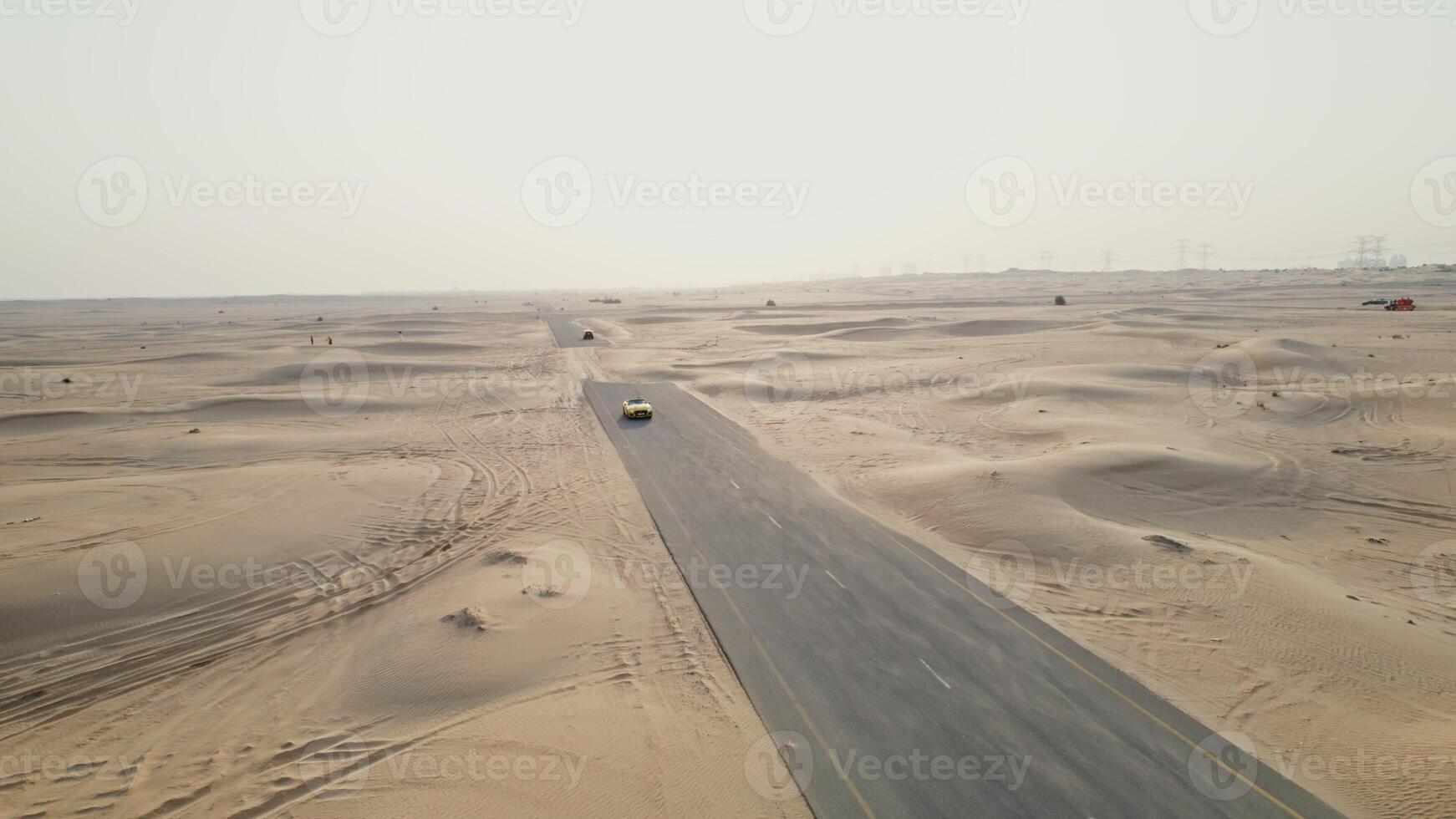 A yellow car is driving along an asphalt road in the desert, raising sand dust behind it. Drone photo
