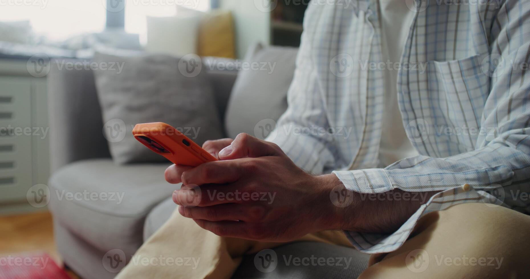 A man in casual clothes smiles while typing on a mobile phone while sitting on the couch at home. The moves from the man's hands to his face photo