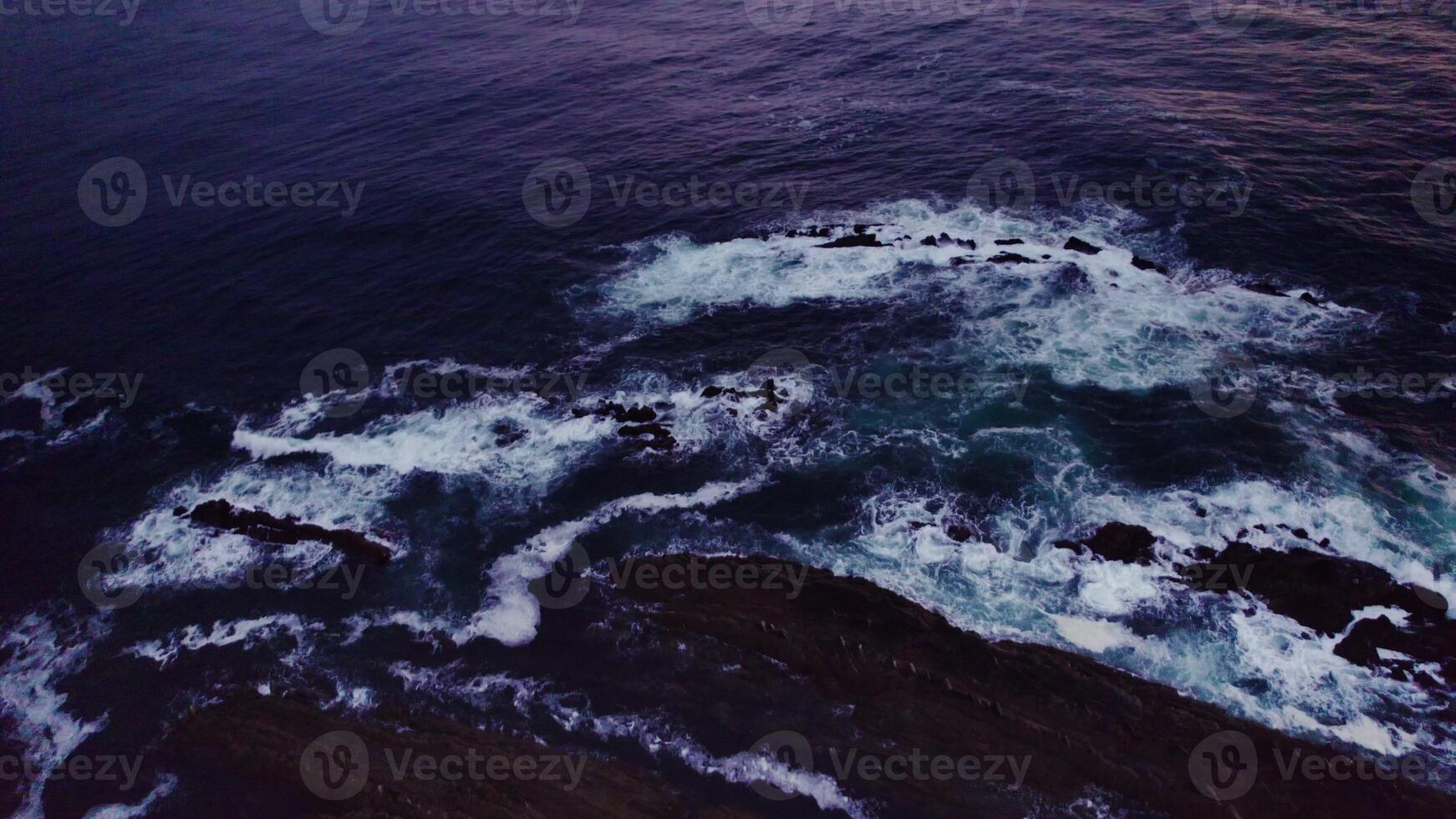 A bird's-eye view of the sea at sunset. Foam waves wash the stone reefs on the shore. Drone photo