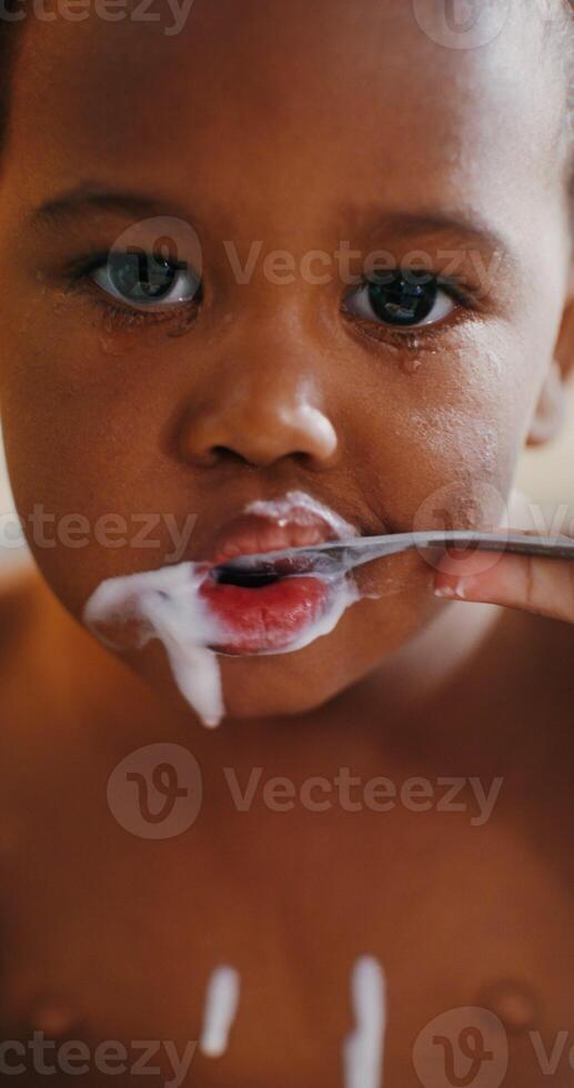 A baby of African-American appearance is eating yogurt with a spoon sitting in a chair, drops of yogurt dripping on her naked tummy photo