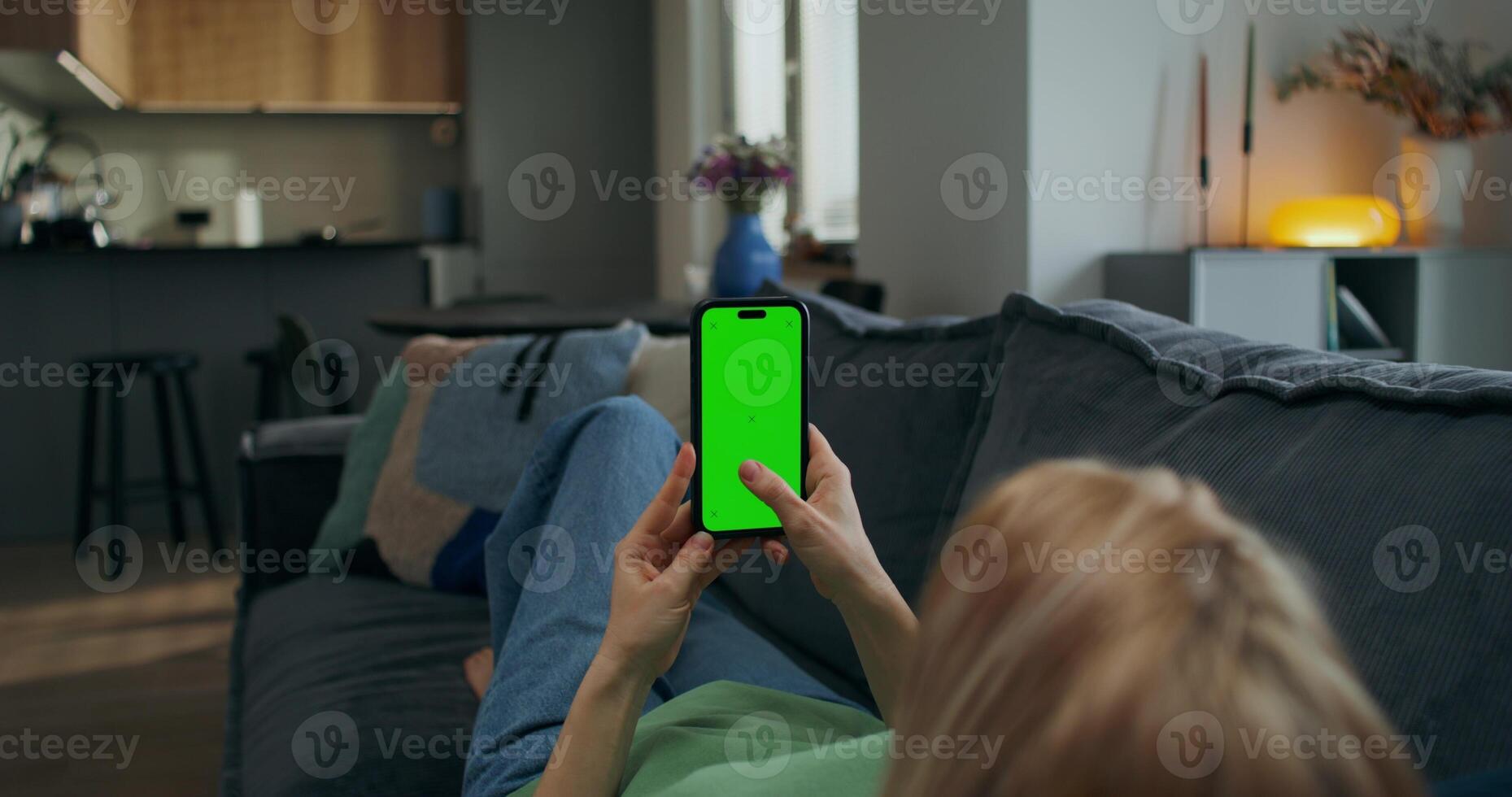 A woman uses a mobile phone with green screen while lying on the couch at home. A rear view of her hands, an unrecognizable person - unrecognizable photo