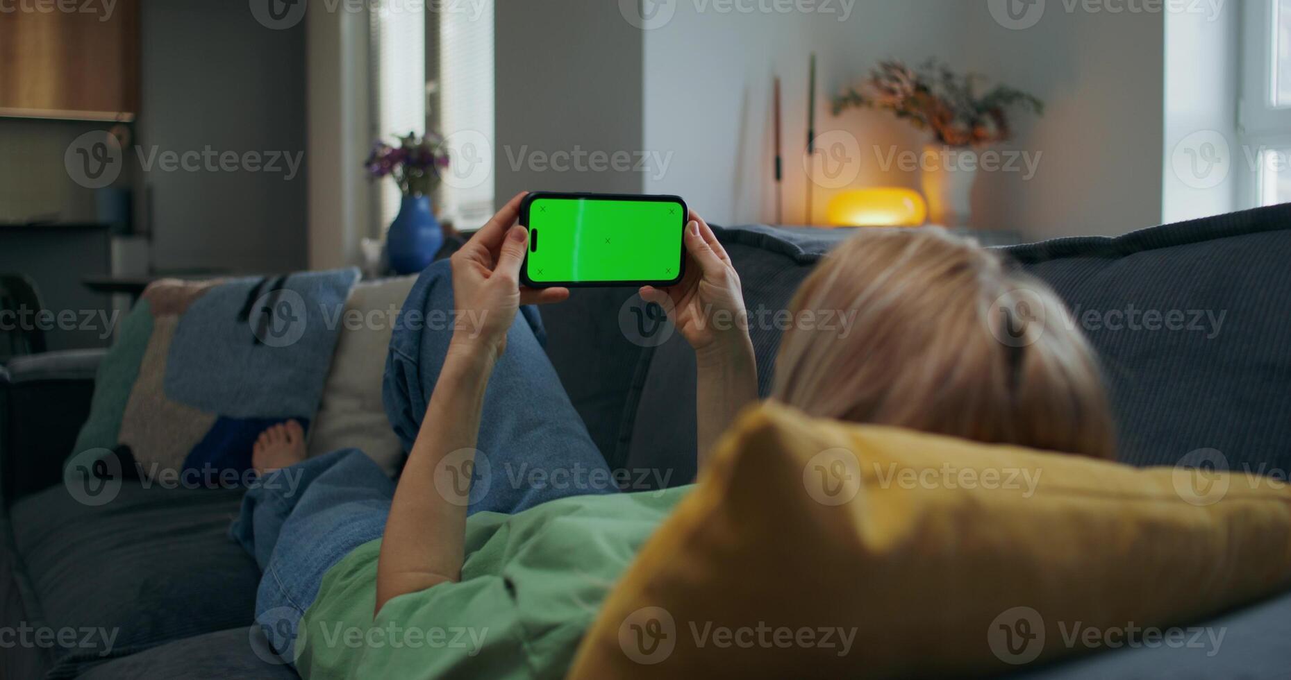 A woman holds a mobile phone with green screen in her hand. She is lying on the couch at home, unrecognizable person photo