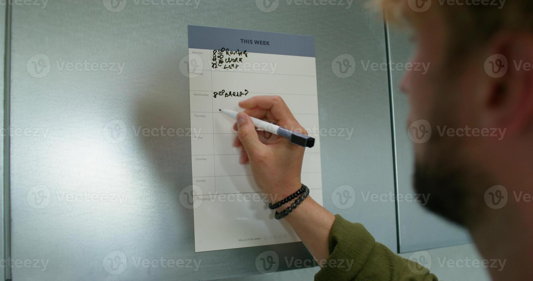 A man in casual clothes fills out a chart with a schedule hanging on the kitchen cabinet. Daylight, home interior - schedule photo