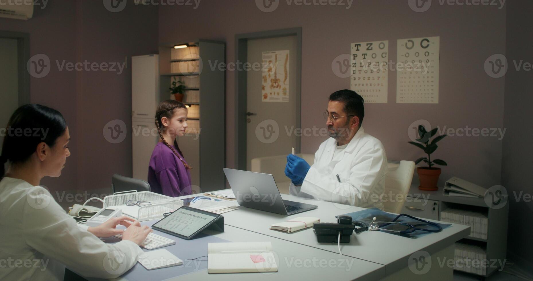 The pediatrician examines the child's throat sitting in the medical office at the clinic. The nurse enters the data into the tablet sitting in front of the doctor - throat photo