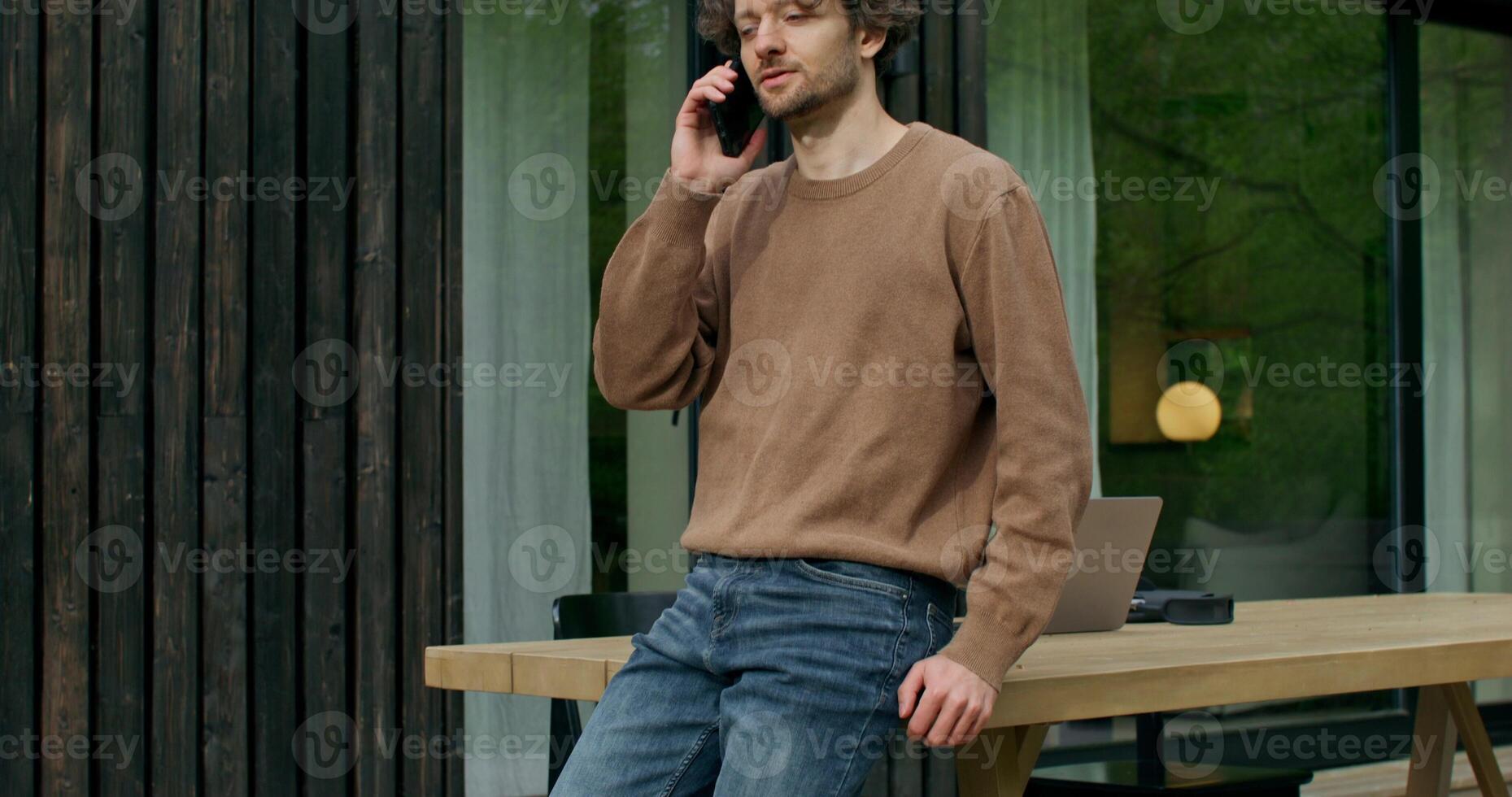 A man in casual clothes is talking on a mobile phone standing on the terrace of a stylish cottage on a summer day. An open laptop is on the table photo