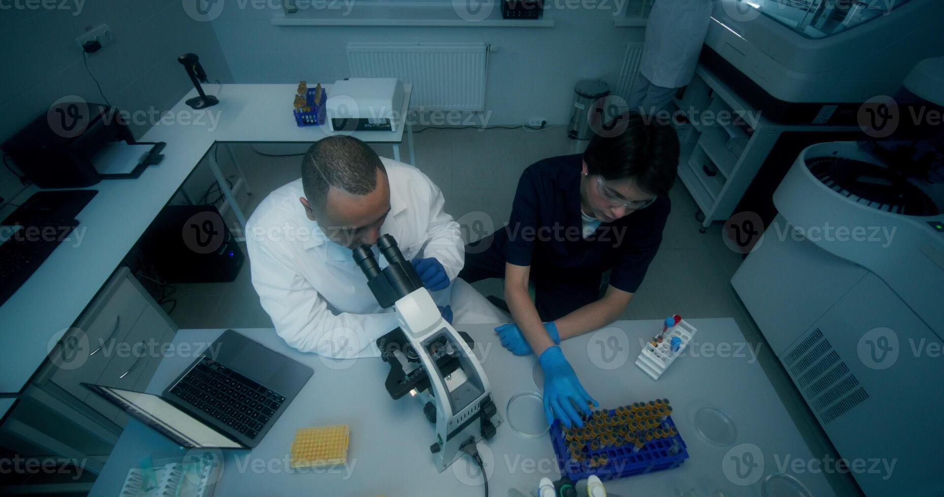 A man and a woman in lab coats are working in a medical laboratory, conducting a blood test and using a microscope. They are talking sitting at the table - medical photo