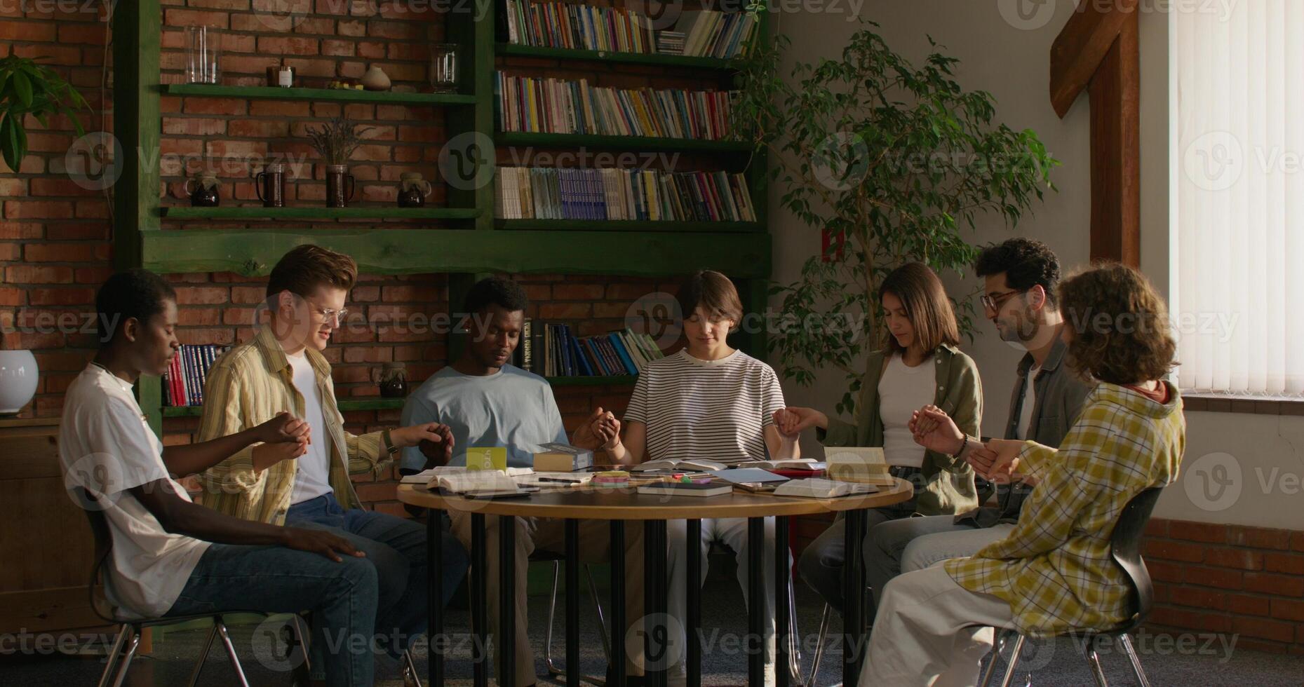 Young people of different nationalities pray holding hands, sitting at a table indoors. Daylight - holding photo