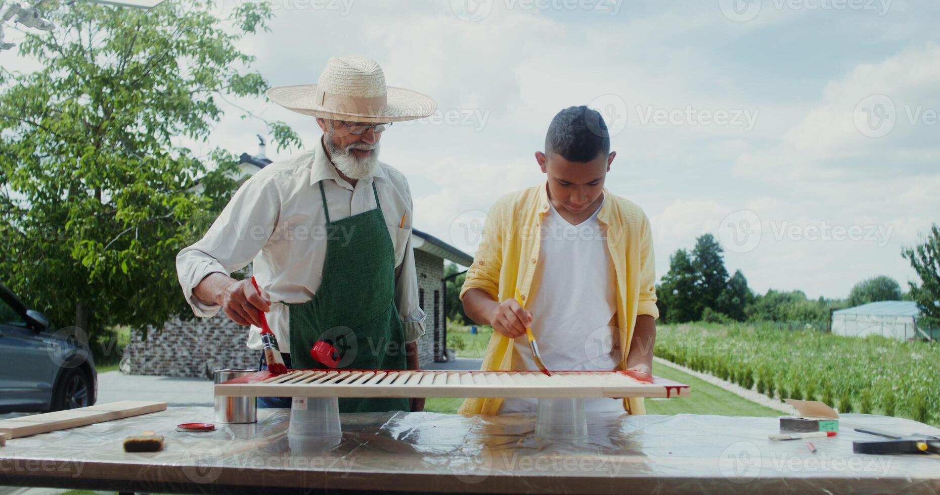 An elderly man in an apron and hat teaches a boy how to paint a door properly while standing in the backyard - standing photo