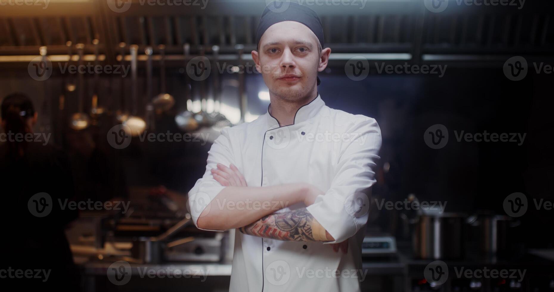 A man in a chef's uniform with a headband looks at the camera with a slight smile, standing in a modern kitchen with his arms crossed at his chest - camera photo