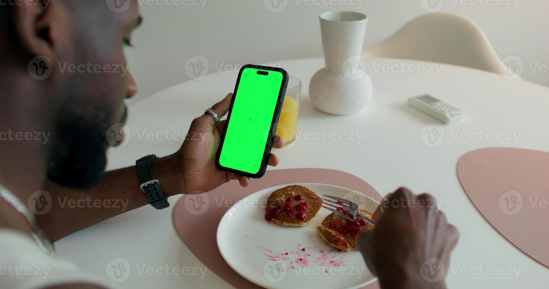 A man eats pancakes and uses a mobile phone with a green screen sitting at a table at home. Daylight photo