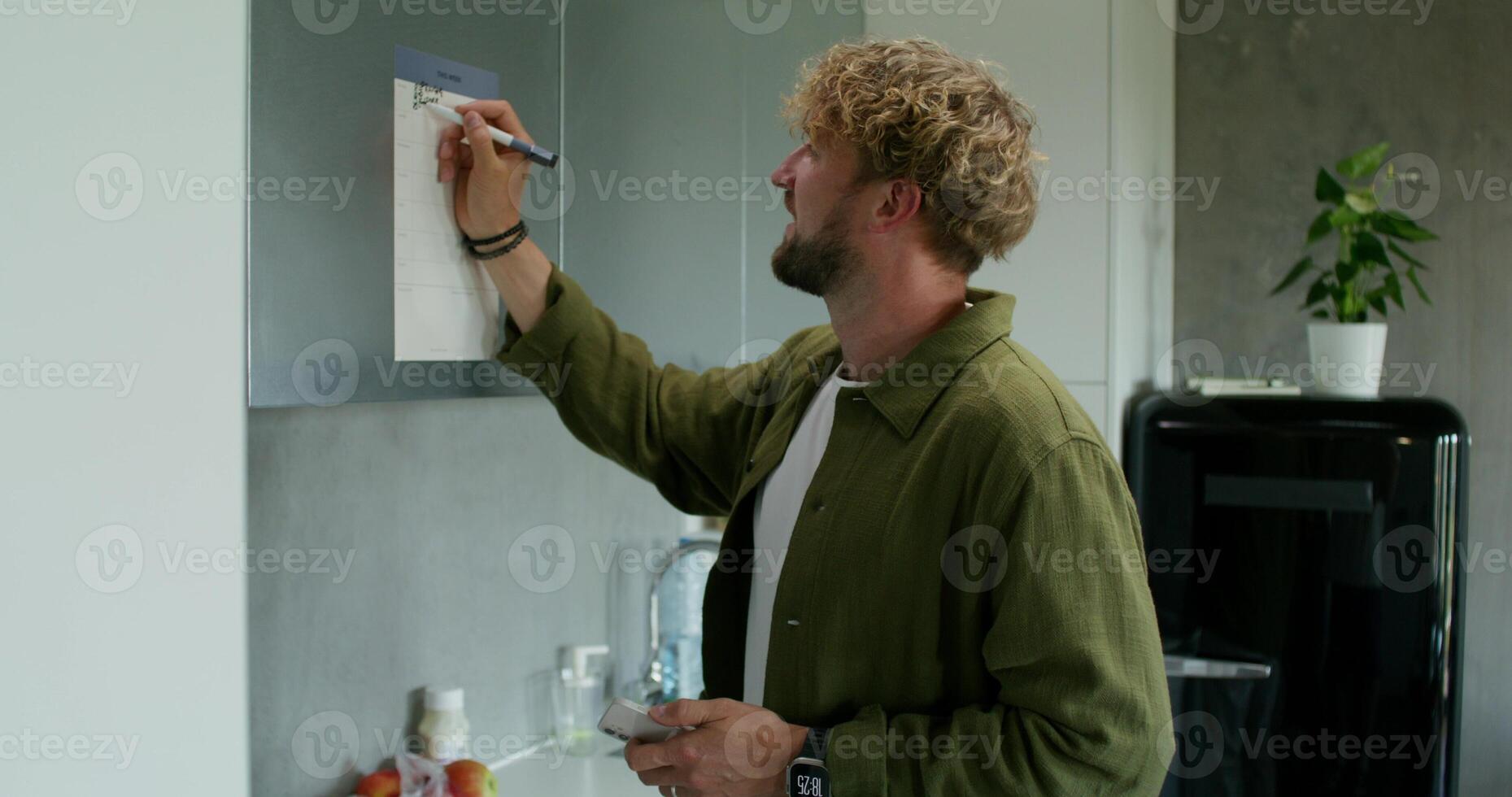 A man in casual clothes fills out a chart with a schedule hanging on the kitchen cabinet. Daylight, home interior - hanging photo