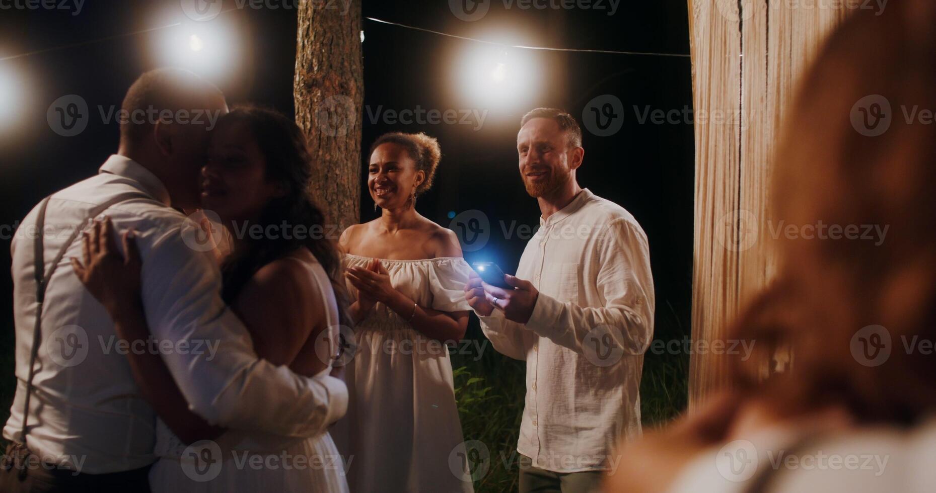 The bride and groom are dancing surrounded by their friends at an outdoor wedding feast at night - friends photo