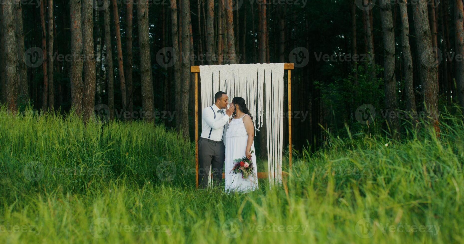 The bride and groom kissing standing near a decorated arch in the forest. Wedding photo shoot - decorated