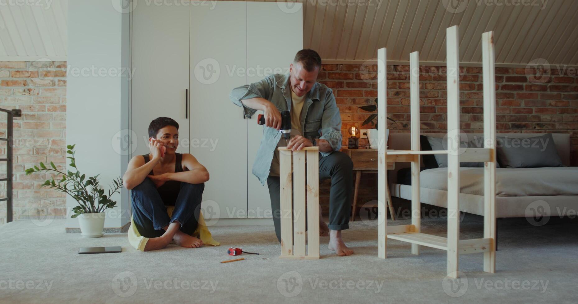 A woman is talking on a mobile phone sitting on the floor next to a man assembling a rack with a screwdriver, they are talking - sitting photo