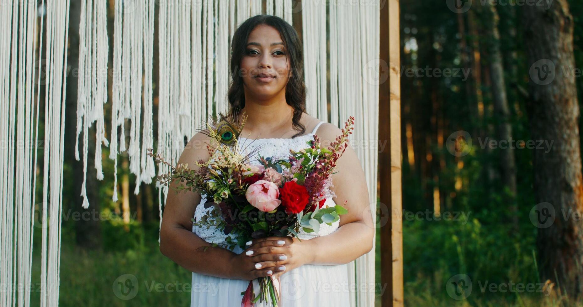 An African-American bride with a bouquet in her hands stands at a decorated arch in the forest and smiles looking at the camera photo