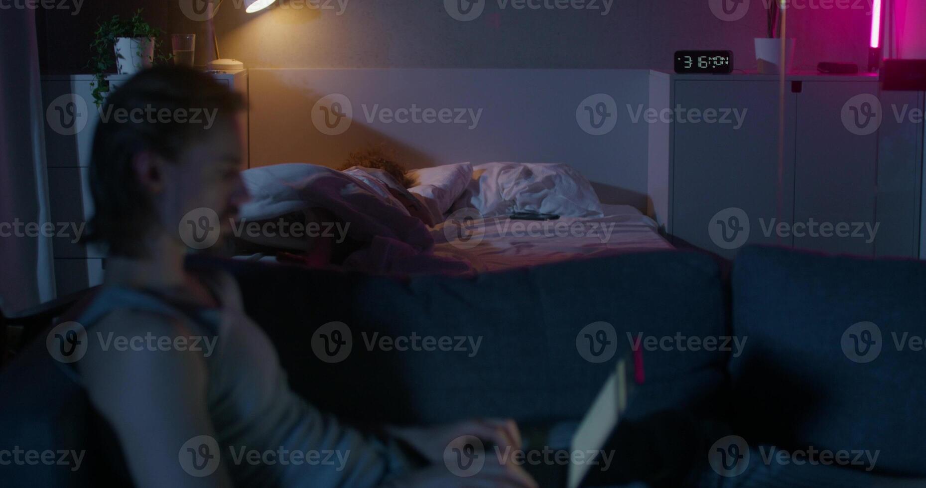 A man uses a laptop sitting on the couch at night. A woman is sleeping in a bed. Dark room, home interior - interior photo
