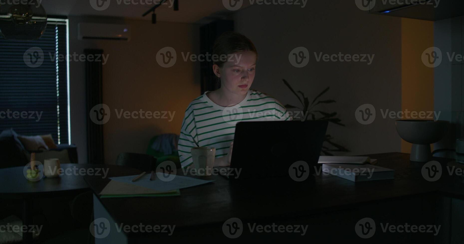A young woman uses a laptop while sitting at a table in the dark. Notebooks and books on the table. Preparing for the session at night - Notebooks photo